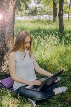 On the banner, a young girl works with a laptop in the fresh air in the park, sitting on the lawn. The concept of remote work. Work as a freelancer. The girl takes courses on a laptop and smiles