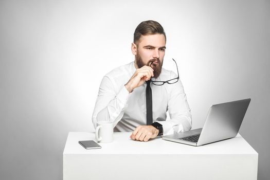 Thoughful bearded young boss in white shirt and black tie are sitting in office on desk and watching daily report on laptop, having new idea and planning own strategy, taking off glasses. Indoor