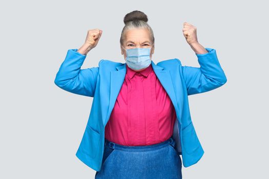 Yes! Happiness success woman with surgical medical mask rejoicing win. Grandmother with light blue suit and pink shirt standing with collected bun hair. indoor studio shot, isolated on gray background