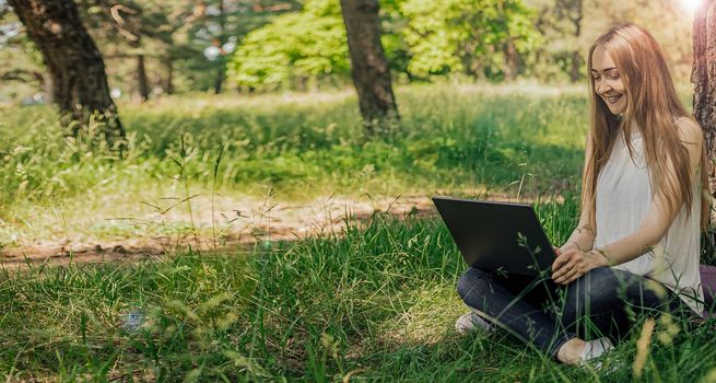 On the banner, a young girl works with a laptop in the fresh air in the park, sitting on the lawn. The concept of remote work. Work as a freelancer. The girl takes courses on a laptop and smiles