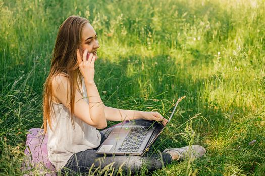 girl sits on the grass and works at a laptop. talking on the phone. freelance. selfeducation. the concept of remote learning and outdoor work