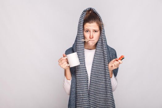 Young woman with gray scarf, holding cup, thermometer and pills. Indoor shot.