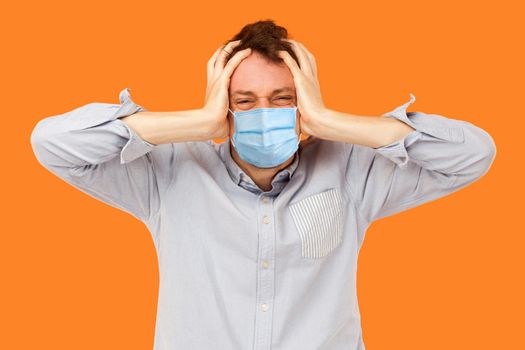 Headache or confusion. Portrait of sad or angry young worker man with surgical medical mask standing and holding his painful head, thinking or enduring. indoor studio shot isolated, orange background.