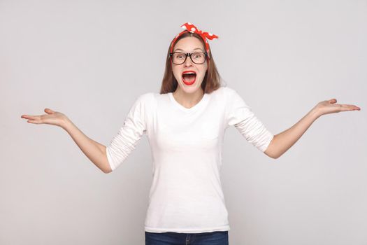 surprised face with raised arms, portrait of beautiful emotional young woman in white t-shirt with freckles, black glasses, red lips and head band. indoor shot, isolated on light gray background.