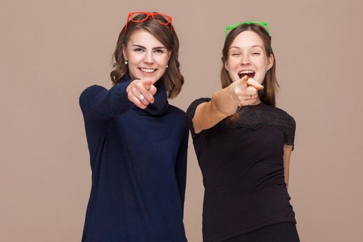 Women standing near each other and pointing fingers at camera. Studio shot, light brown background