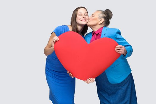 Grandmother kissing granddaughter standing, holding big heart shape, looking at camera with toothy smile. Big love in family. Relations in the family. indoor studio shot, isolated on gray background