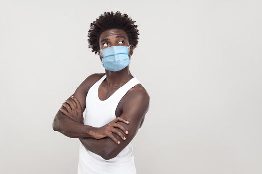 Portrait of thoughtful young man wearing white shirt with surgical medical mask standing, folding arms and looking away and thinking what to do, boring. indoor studio shot isolated on gray background.