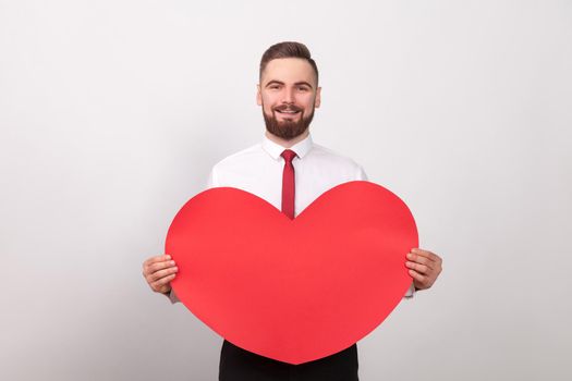 Valentines day. Energy businessman toothy smiling, holding big red heart. Indoor, studio shot, isolated on gray background