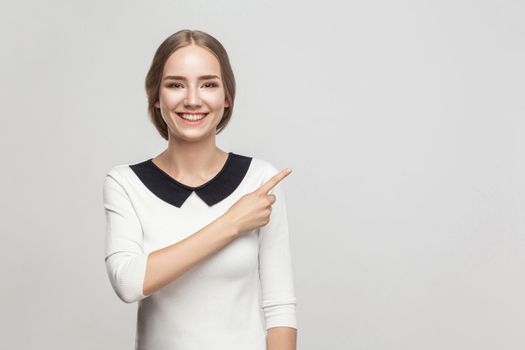 Business woman pointing finger at copy space, toothy smiling and looking at camera. Studio shot, gray background