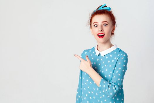 Excited young redhead caucasian girl with hair knot pointing her index finger sideways, raising eyebrows and keeping mouth wide opened, showing something surprising on grey copy space wall.