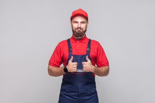 Work are done! Portrait of young satisfied cheerful repairman with beard in blue overall, red t-shirt and cap, standing and showing thumps up with smile. Grey background, indoor studio shot isolated.