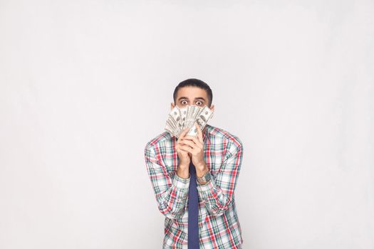 Portrait of rich handsome young adult businessman in colorful checkered shirt with blue tie standing and showing fan of dollars with big crazy eyes. Indoor, studio shot, isolated on grey background.