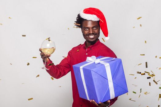 Around happiness afro man in red cap, flies gold metaphane, man holding champagne glass and gift box, looking at camera and toothy smiling. Studio shot. Gray background