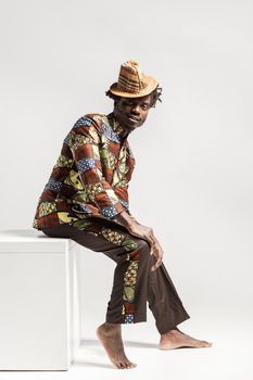 Happy fashion african man in traditional clothes sit on cube . Indoor, isolated on gray background
