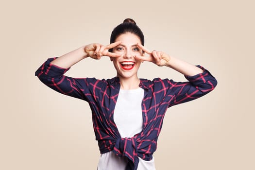 Victory and peace sign. Happy toothy smiley young woman holding hand on eyes and showing victory sign with satisfied face. studio shot on beige background.