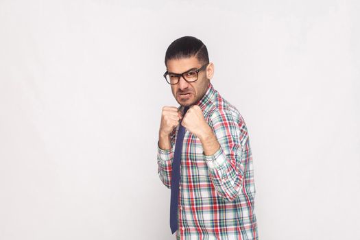 Angry bearded businessman in colorful checkered shirt, blue tie and black eyeglasses standing and looking at camera with aggressive face and fist. indoor studio shot, isolated on light grey background