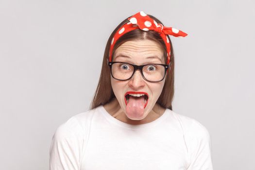 tongue out of surprised, or shocked crazy emotional young woman in white t-shirt with freckles, black glasses, red lips and head band. indoor studio shot, isolated on light gray background.