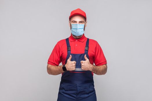 Work are done! Portrait of young man with surgical medical mask in blue overall, red t-shirt , cap, standing and showing thumps up and looking at camera. Grey background, indoor studio shot isolated.