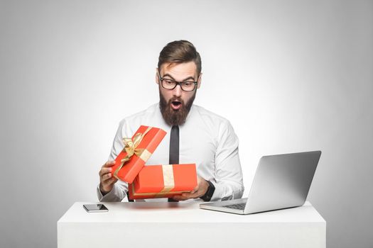 Wow! Suprised young manager in white shirt and black tie are sitting in office and unboxing present with shocked face, big eyes and opened mouth. Indoor, isolated, studio shot, gray background