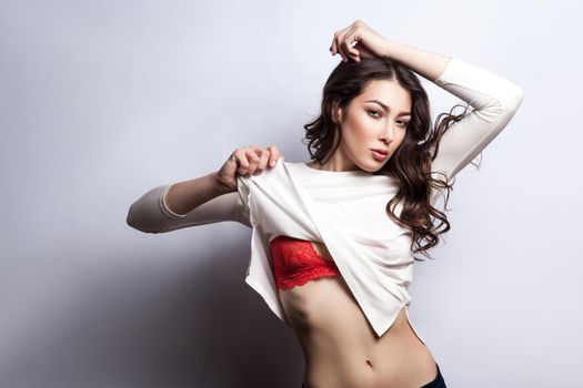 Beautiful young sexy fashion model posing in bra and undressing her white t shirt and looking at camera with passion. Indoor, studio shot on gray background