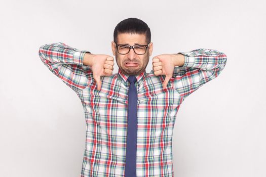 Sad unhappy bearded businessman in colorful checkered shirt, blue tie and eyeglasses standing and looking with thumbs up and dislike gesture. indoor studio shot, isolated on light grey background.