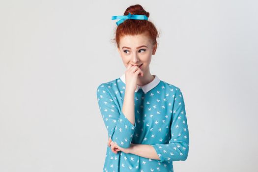 People, youth and tenderness concept. Portrait of young redhead female model having shy cute smile, holding hand on her lips, posing indoors. Isolated studio shot on gray background.