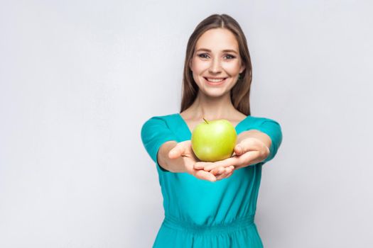 Young beautiful woman with freckles and green dress holding apple and sharing with smile. studio shot, isolated on light gray background.