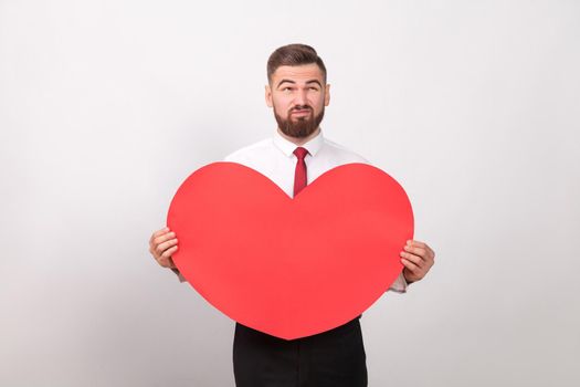 Bearded man thoughtful, holding red heart and thinking of love. Indoor, studio shot, isolated on gray background