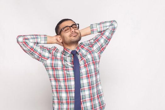 Portrait of dreamy handsome bearded businessman in checkered shirt, blue tie and eyeglasses standing with hand behind head and smiling with closed eyes. indoor studio shot, isolated on gray background