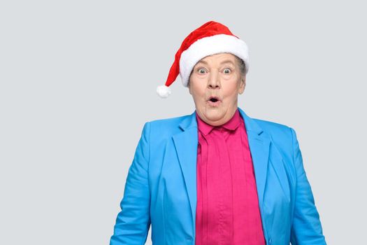 Surprised grandmother in colorful casual style with blue suit and christmas santa red cap standing and looking at camera with amazed face and open mouth and eyes. indoor, isolated on gray background