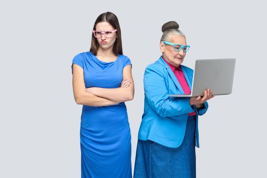 Unhappy confused granddaughter standing near grandmother holding and working on her laptop and making conflict. relationship or mutual understanding. indoor studio shot, isolated on gray background