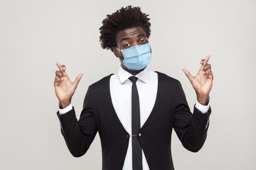 Hopeful or wishful. Portrait of worry young handsome worker man wearing black suit with surgical medical mask standing with crossed fingers and hope. indoor studio shot isolated on gray background.