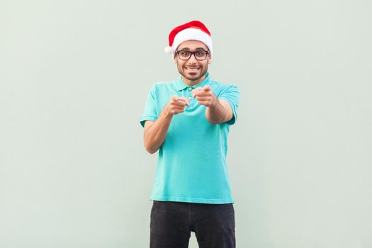 Hey you! Its christmas man! Bearded man in santa hat and glasses, pointing finger and looking at camera with toothy smile. On gray background. Indoor, studio shot