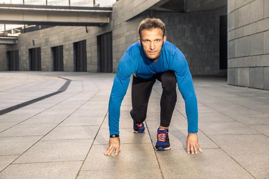 Handsome middle aged serious man in blue black sports uniform and headphones and fitness tracker is running in the city.