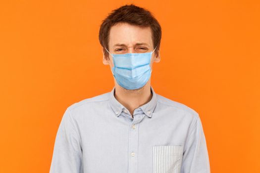 Portrait of sad alone young worker man with surgical medical mask standing and looking at camera with upset frown face. indoor studio shot isolated on orange background.