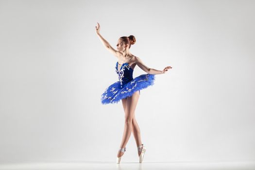 attractive ballerina with bun collected hair wearing blue dress and pointe shoes performing in white studio. indoor, studio shot.