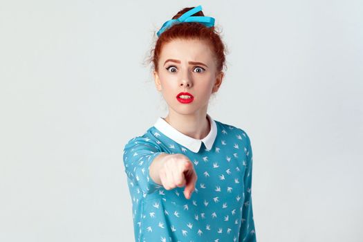 The beautiful caucasian girl, wearing blue dress, opening mouths widely, having surprised shocked looks, pointing finger at camera. Selective focus. Isolated studio shot on gray background