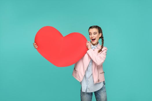 Amazed beautiful girl in casual style, pigtail hairs and jacket, standing and holding and looking at big red heart shape with surprised face. indoor, studio shot isolated on blue or green background