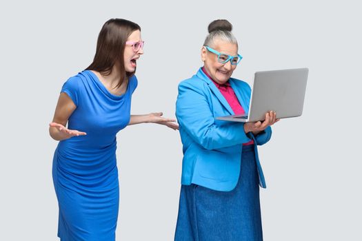 Unhappy young woman in blue dress shouting at aged woman working on laptop. Relations or relationship on family between granddaughter and grandmother. indoor, studio shot, isolated on gray background