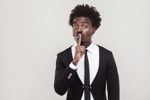 Secret or quiet concept. African businessman showing shh sign at camera. Indoor shot, gray background