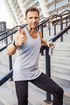 Happy smily sportsman holding bottle of water, standing on steps looking at camera smiling and showing thumb . Thumbs up, fitness, sport, exercising and people healthy lifestyle concept.