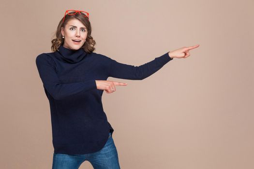 Adult woman in red glasses looking at camera and pointing at copy space. Studio shot, light brown background