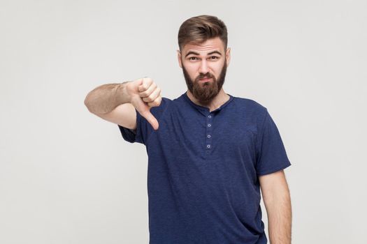 Portrait of unsatisfied bearded man with thumbs down and blue t-shirt against gray background. Studio shot.