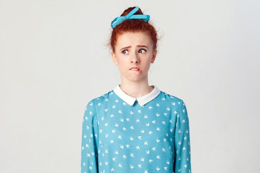 The cute redhead girl looking away, having doubtful and indecisive face expression, pursuing her lips as if forbidden to say anything. Isolated studio shot on gray background.