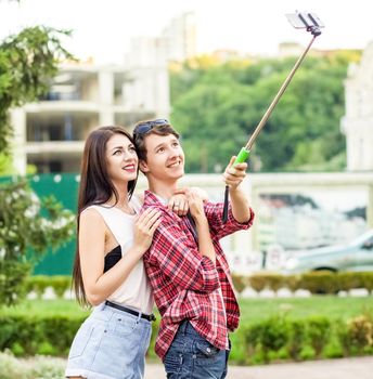 happy young tourists couple taking a selfie with smartphone on the monopod in city. The man is holding the stick and shooting looking at phone with happiness..