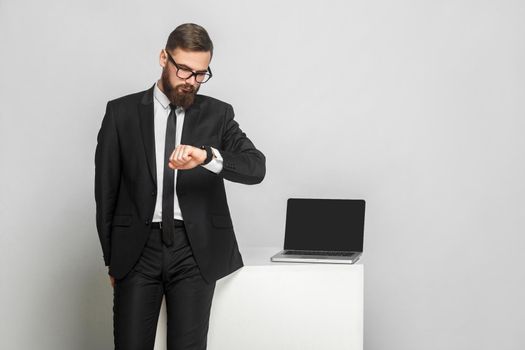 Portrait of handsome thoughful bearded young businessman in corporated format wear in black suit are standing and checking time on his own hand watch. Isolated, studio shot, indoor, gray background