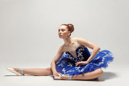 attractive ballerina with bun collected hair wearing blue dress and pointe shoes sitting on white studio floor. indoor, studio shot.