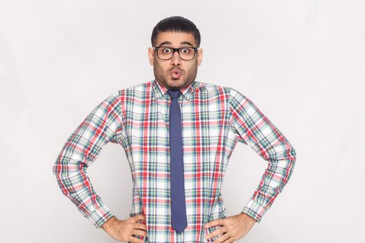 Surprised handsome bearded businessman in checkered shirt, blue tie and black eyeglasses standing with hands on waist and looking at camera with excited face and big eyes. Isolated on grey background