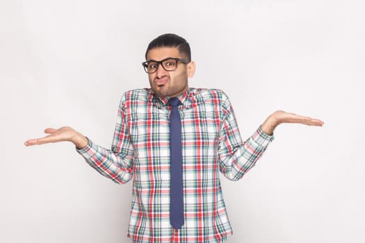 I don't know. confused bearded businessman in checkered shirt, blue tie and eyeglasses standing, raised arms and looking at camera with doubtful face. studio shot, isolated on light grey background.