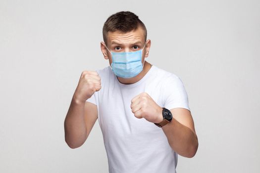 Portrait of serious young man in white shirt with surgical medical mask standing with boxing fists, looking at camera and ready to attack or defence. indoor studio shot, isolated on gray background.